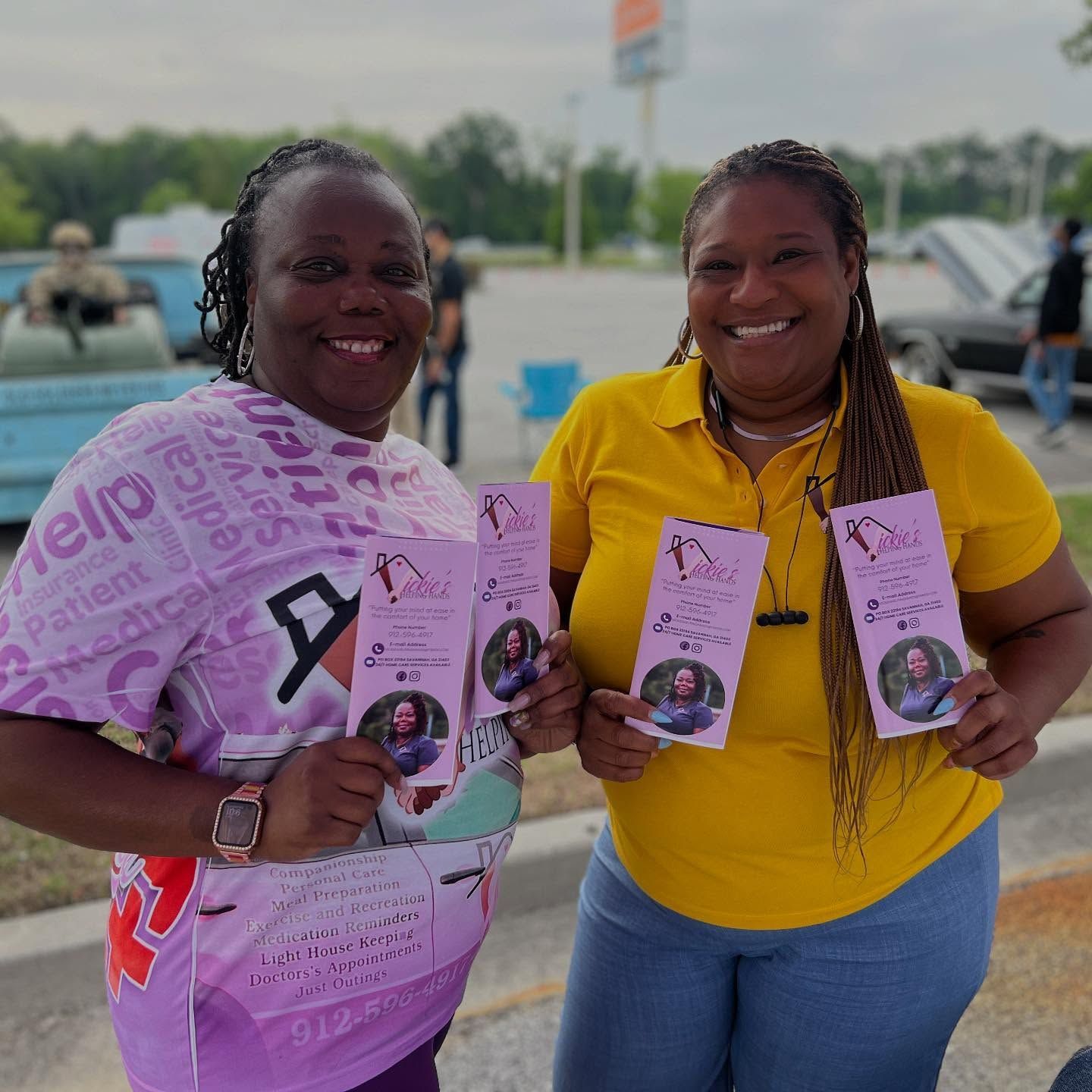 Two people smiling, holding brochures outdoors. One wears a pink shirt, the other yellow. Both are near cars.