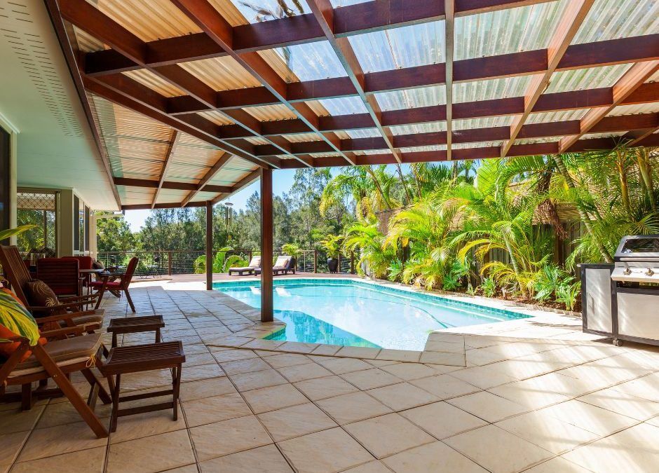 Patio with pool: Wooden chairs, barbecue, tiled floor, pool, and tropical foliage under a latticed roof.
