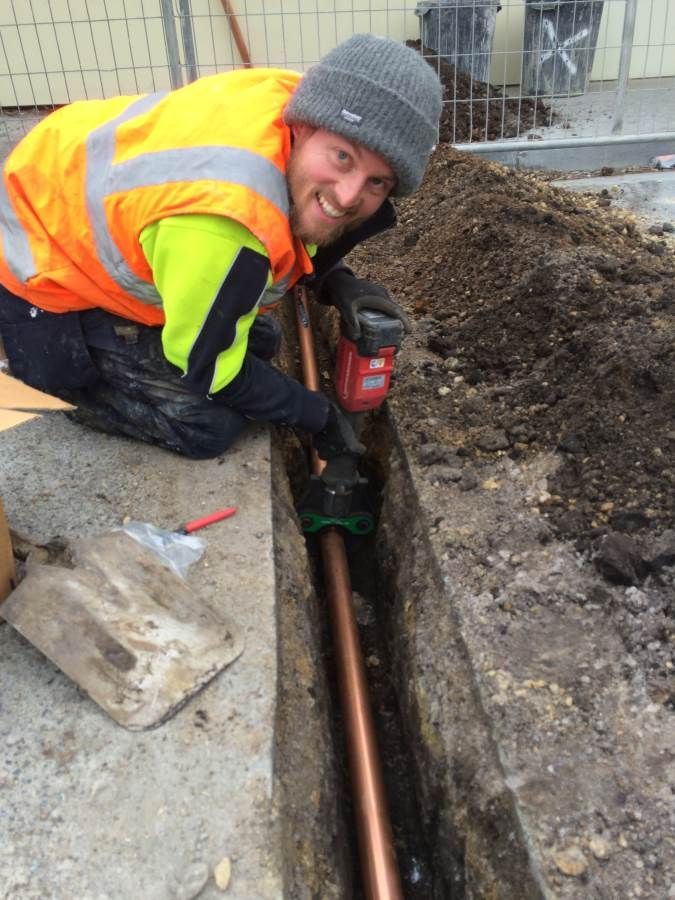 Construction worker installing a copper pipe in a trench, wearing safety vest and beanie, smiling.