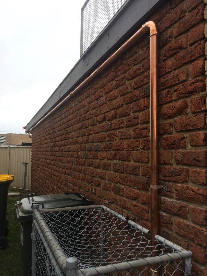 Copper pipe running along a red brick wall, above trash bins and a metal fence.