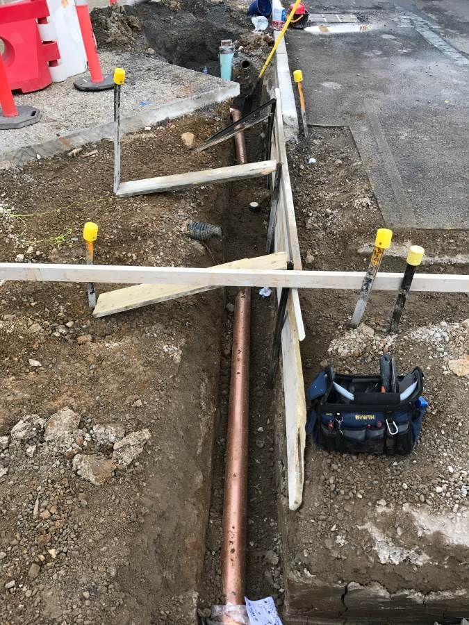 Trench with copper pipe, tools, and wooden supports on a street, likely for utility work.