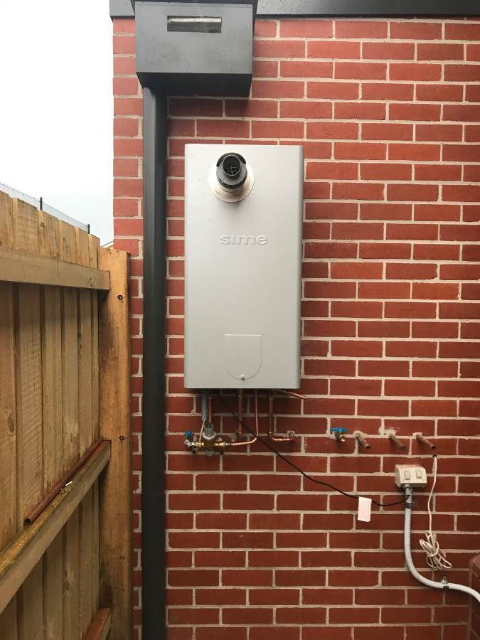 Gas water heater mounted on a red brick wall with exhaust pipe and supply lines.