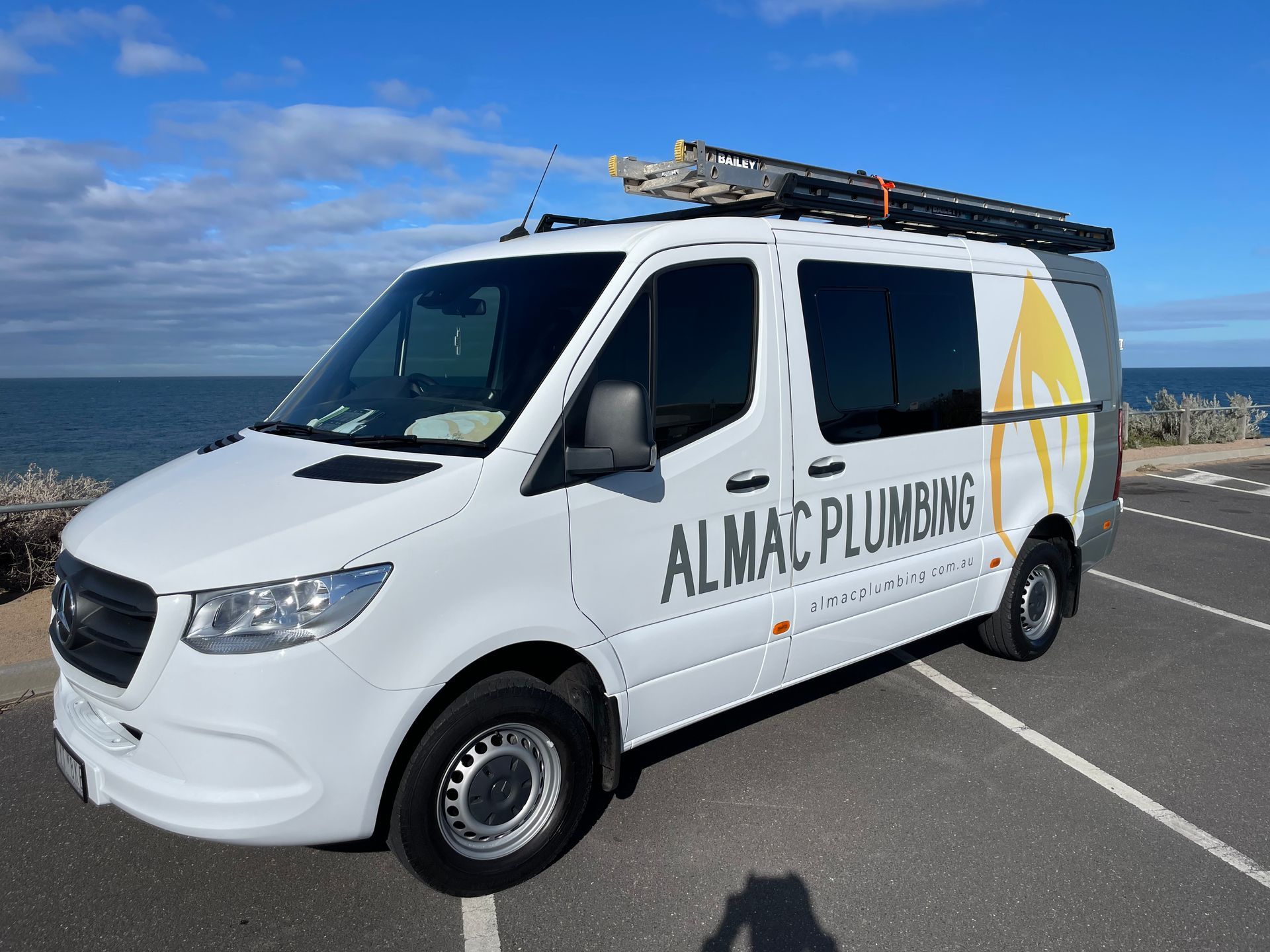 White Almac Plumbing van parked by the ocean with ladder on top.