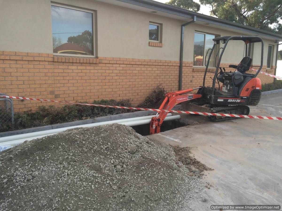 Mini excavator digging near a building's foundation, creating a trench. Gravel pile in foreground. Red, white tape.