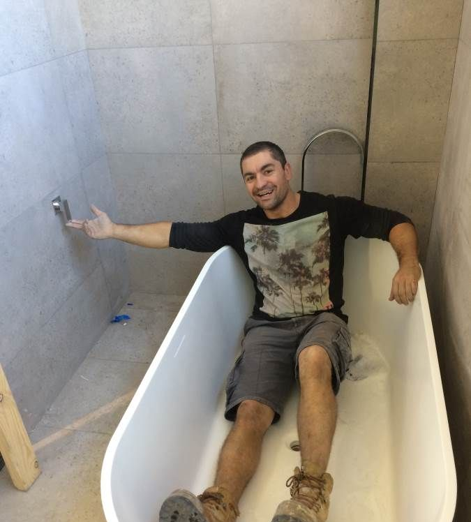 Man sitting in a white bathtub in a bathroom under construction, gesturing toward the faucet.