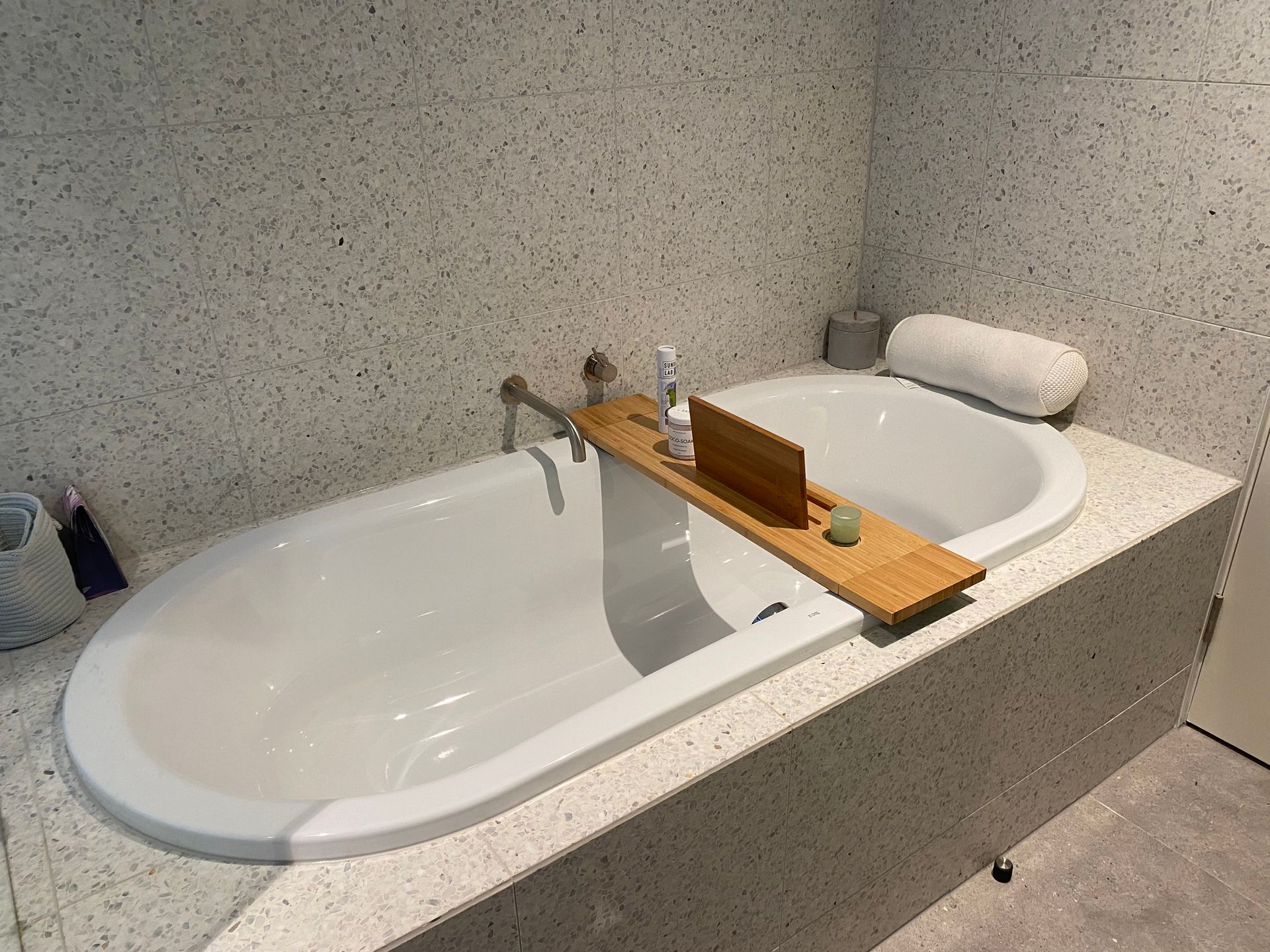 Oval bathtub with a wooden tray holding toiletries. Gray speckled walls and platform. A white pillow rests on the edge.