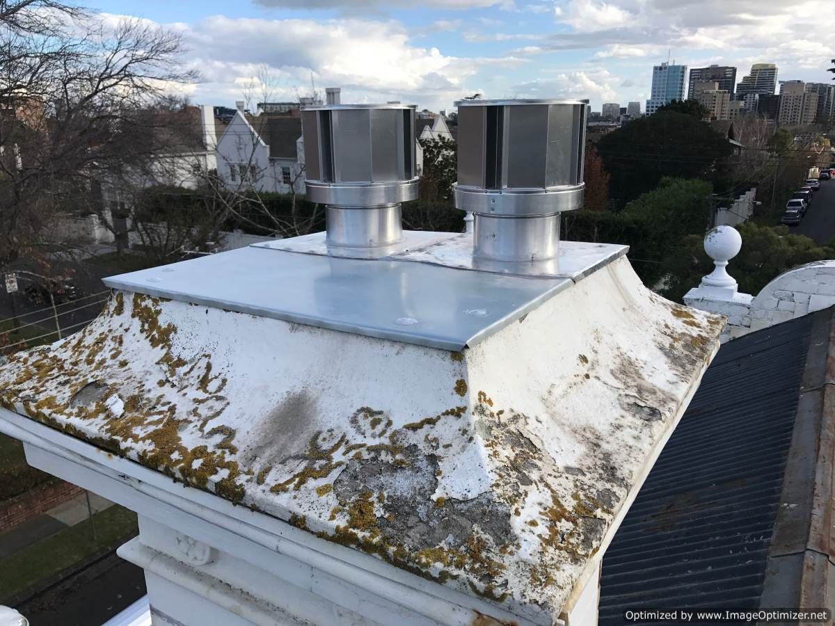 White chimney with two metal vents, mossy edges, city in background.