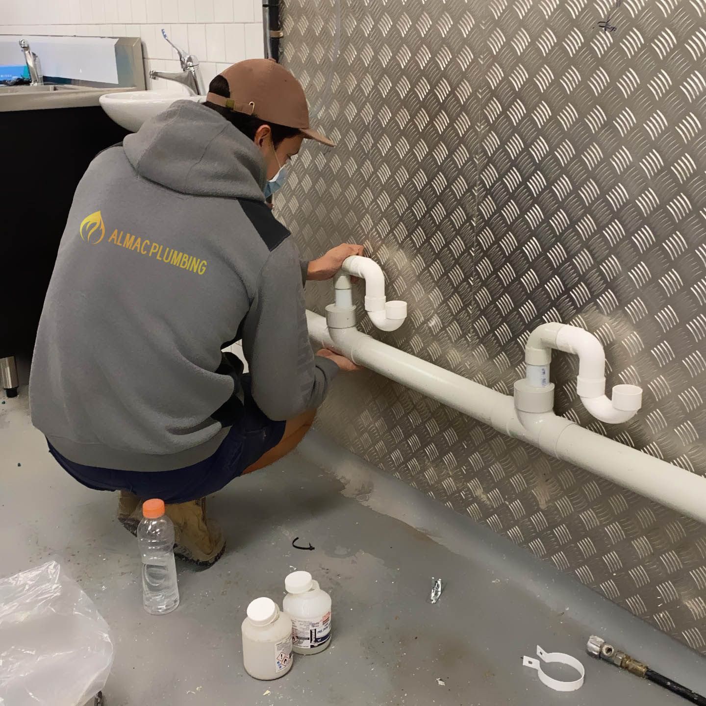 Plumber installing white pipes on a diamond-plate wall, wearing gray hoodie and shorts.
