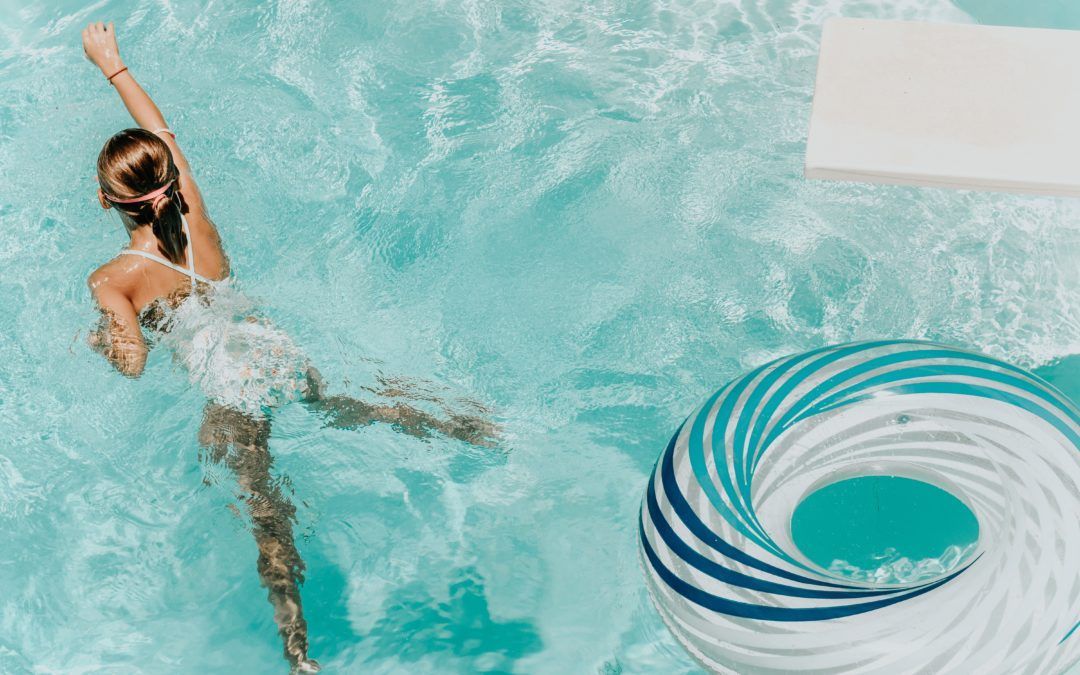 Woman swimming in a clear blue pool, reaching with arm. A striped inflatable ring floats nearby.