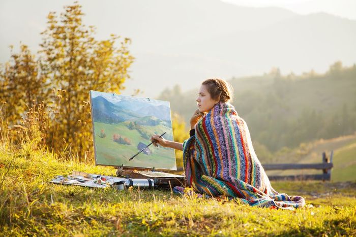 A woman is sitting in the grass painting a landscape.