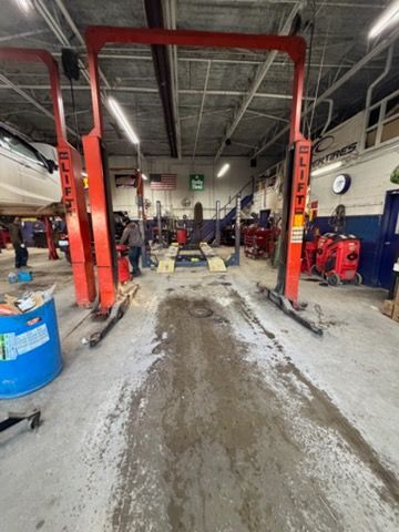 Inside a car repair shop, a two-post lift is centered. Red lifts stand on a concrete floor. A worker stands nearby.
