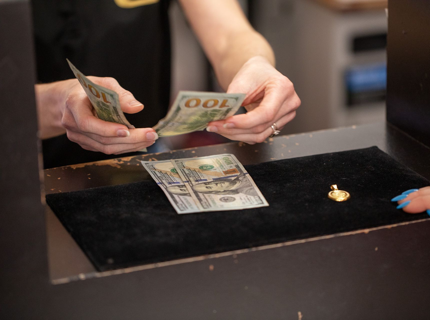 Hands counting $100 bills over a counter. A piece of jewelry sits on a black velvet pad.