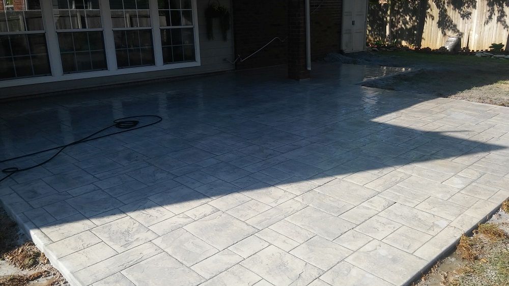 Sunlit stone patio beside a house, with a shadowed backyard and glass doors in the background.