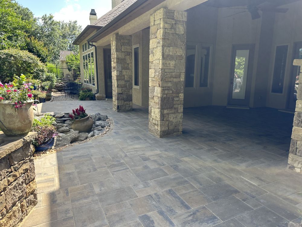 Stone patio with covered porch, columns, and potted plants beside a landscaped garden