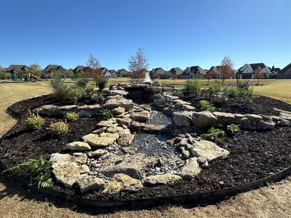 Circular rock garden with layered stones and small shrubs in a grassy suburban park under a blue sky