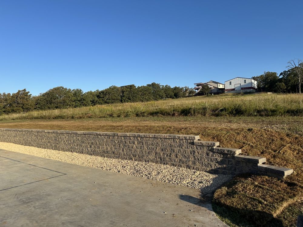 Stone retaining wall beside a paved road, with grassy field and trees under a clear blue sky.