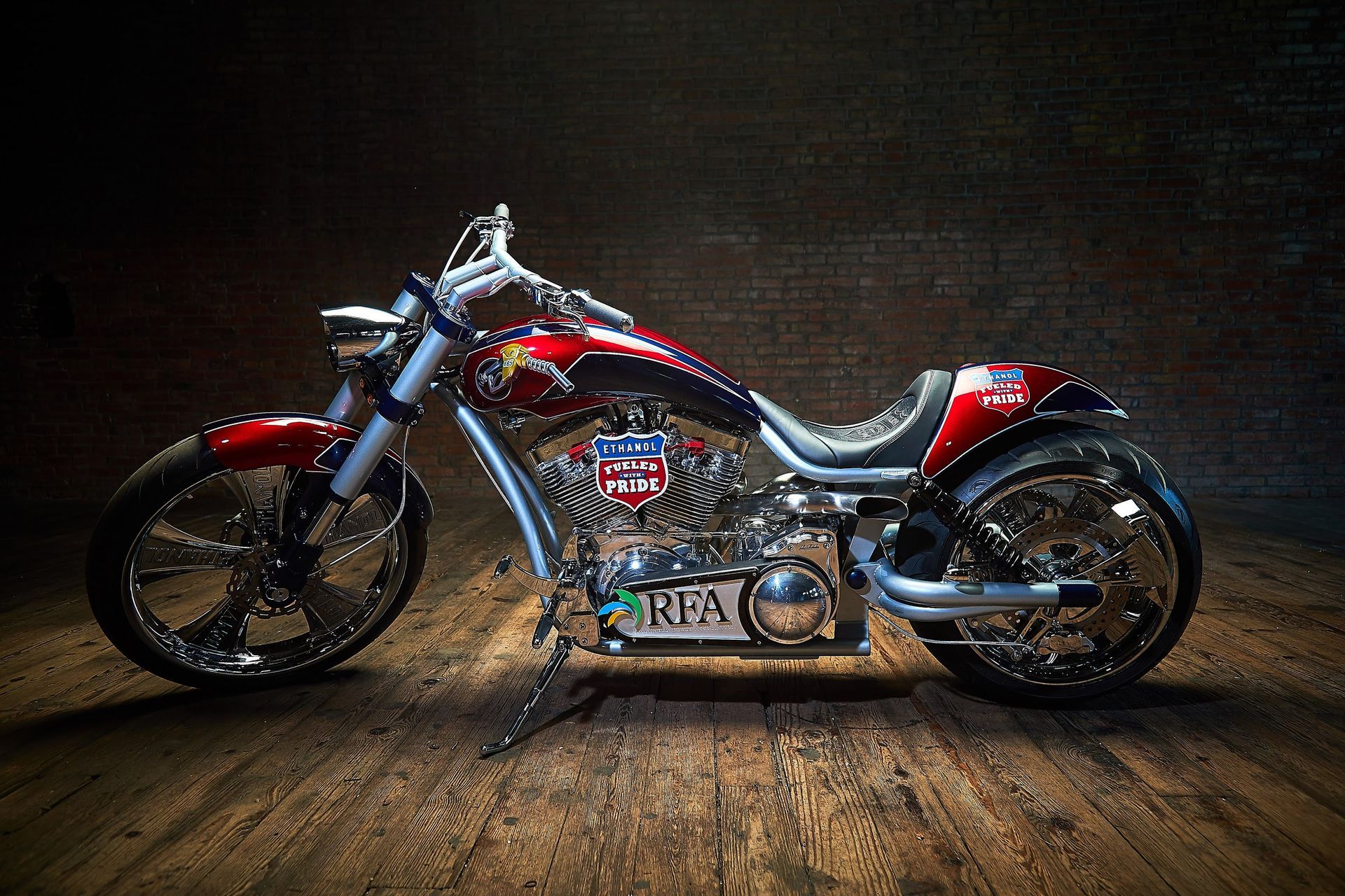 A red and black motorcycle is parked on a wooden floor in front of a brick wall.