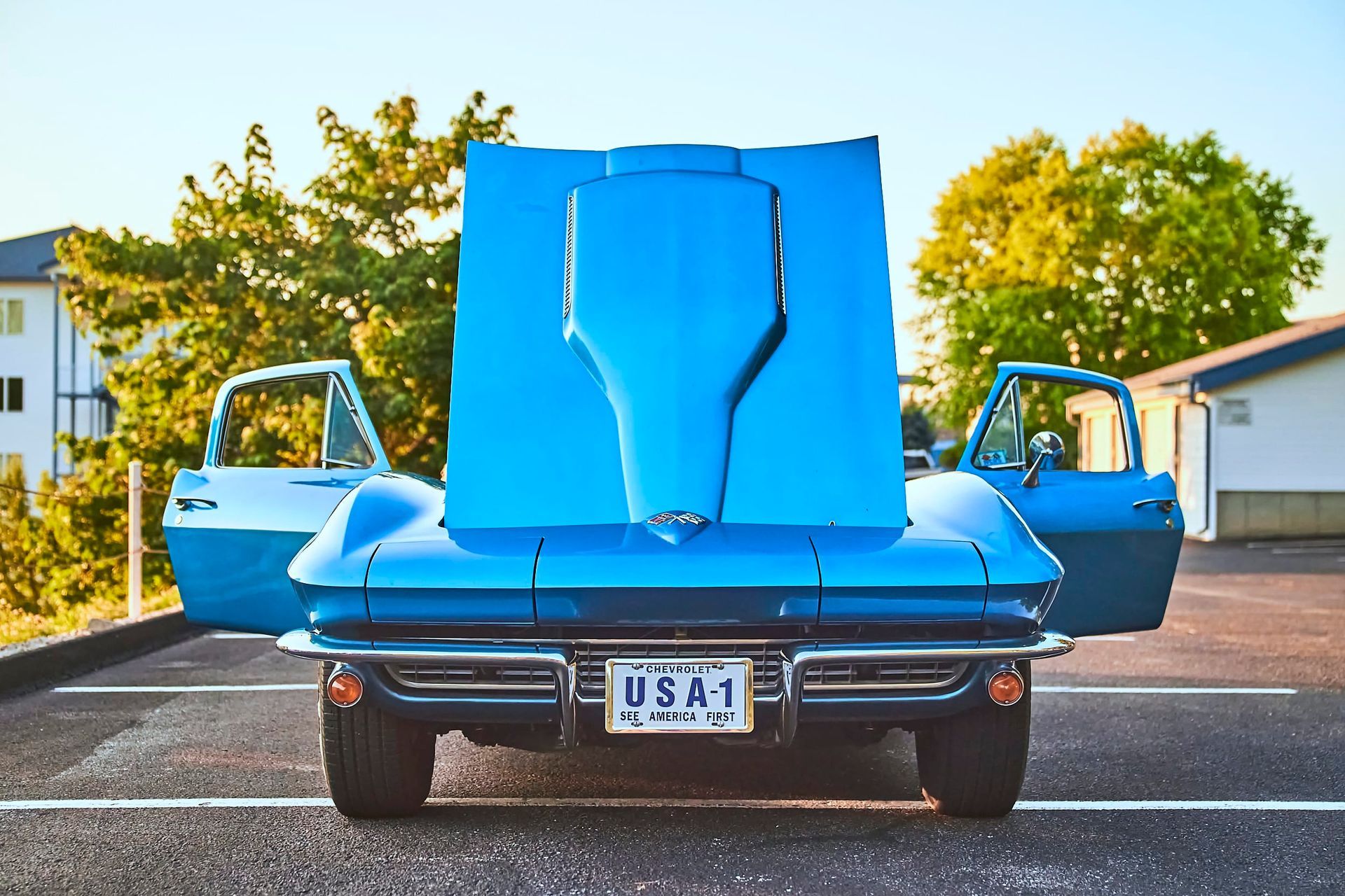 A classic blue Corvette with its hood and doors open, parked on asphalt. The license plate reads