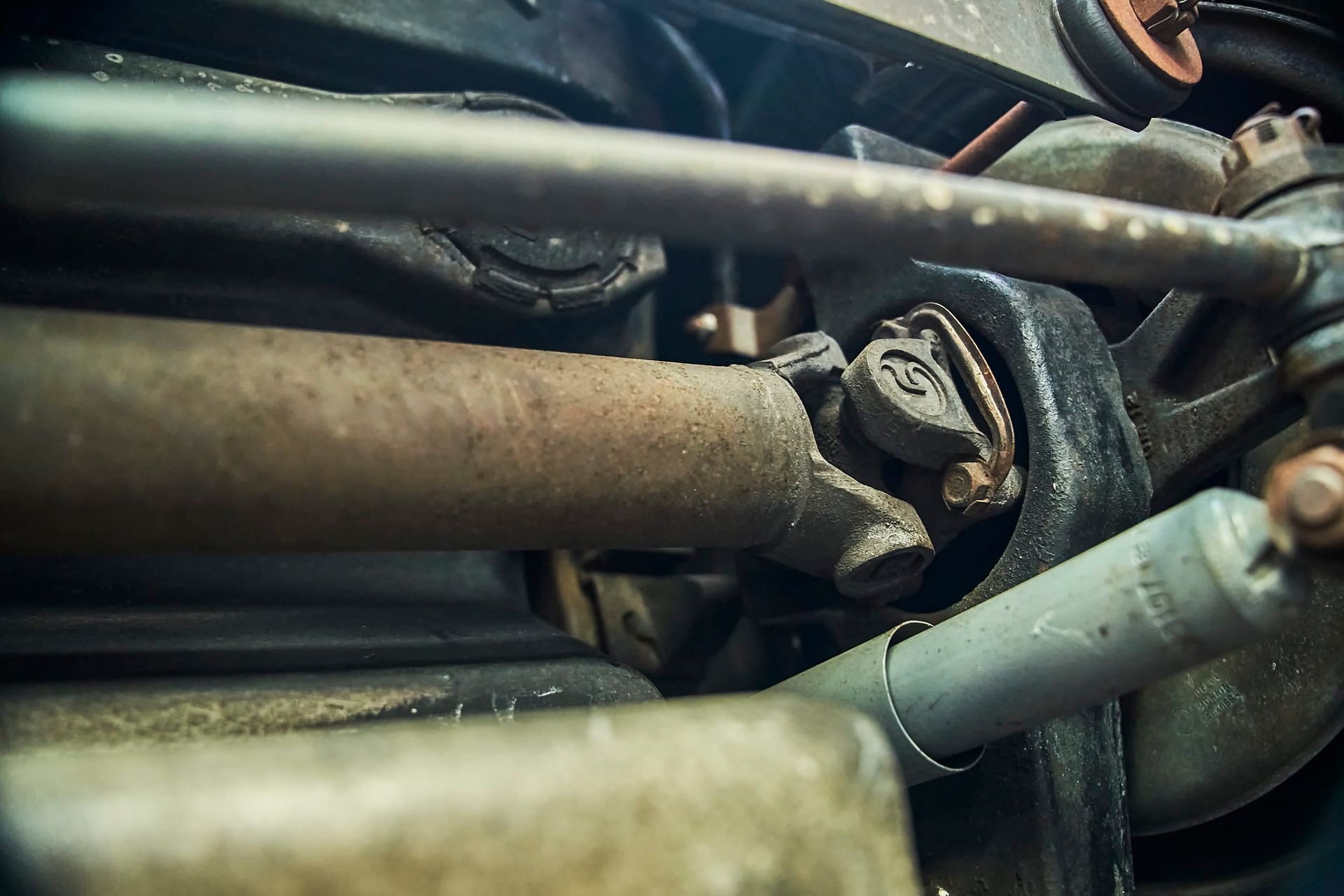 Close-up view of a rusty car driveshaft, showing a U-joint and surrounding components. The shot is from underneath the vehicle.