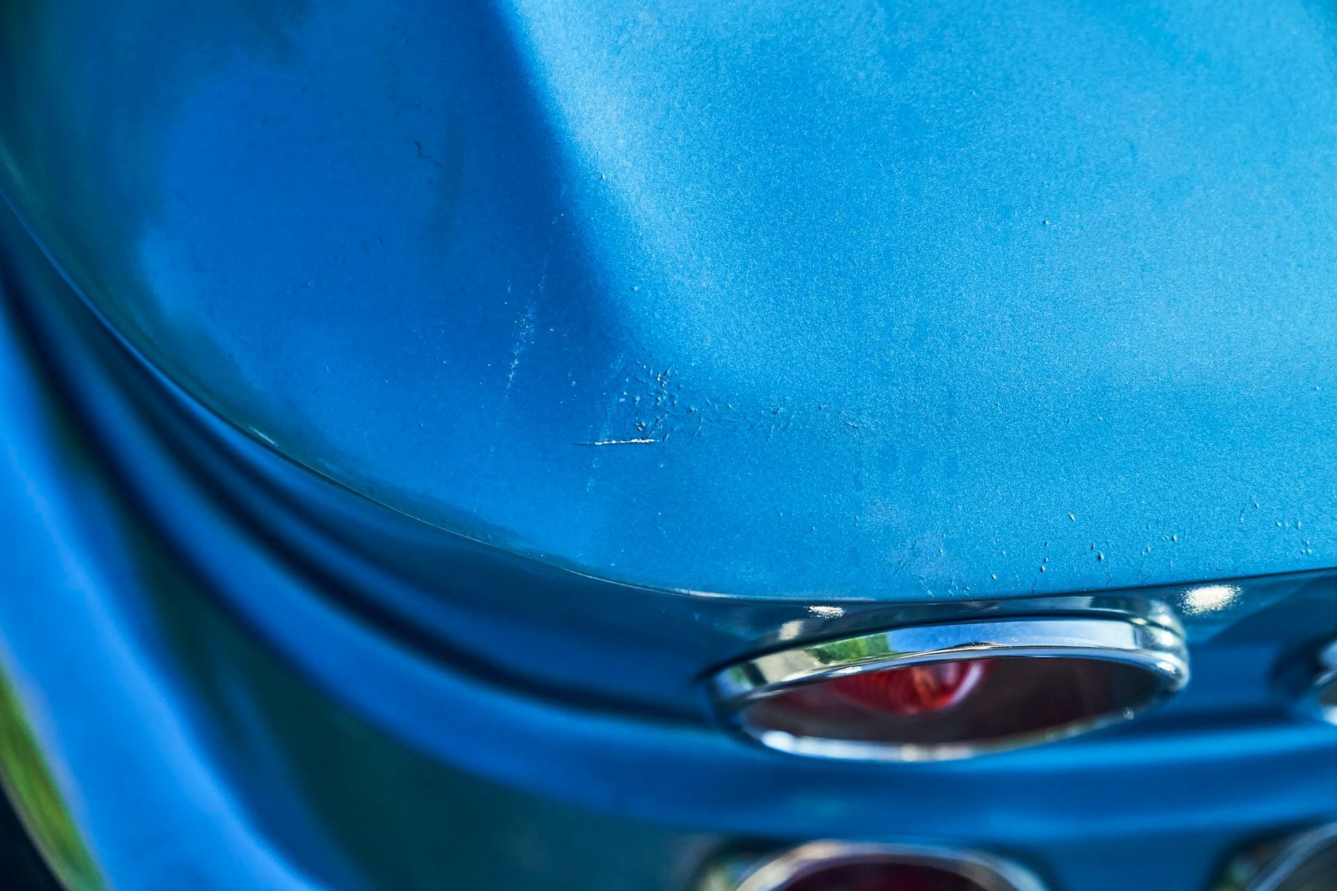 Close-up of a blue car's rounded back end. Small paint chips are visible near the top, above a chrome-ringed taillight.
