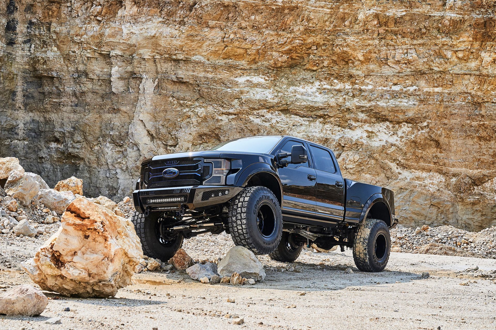 A black truck is parked in the dirt in front of a rock wall.