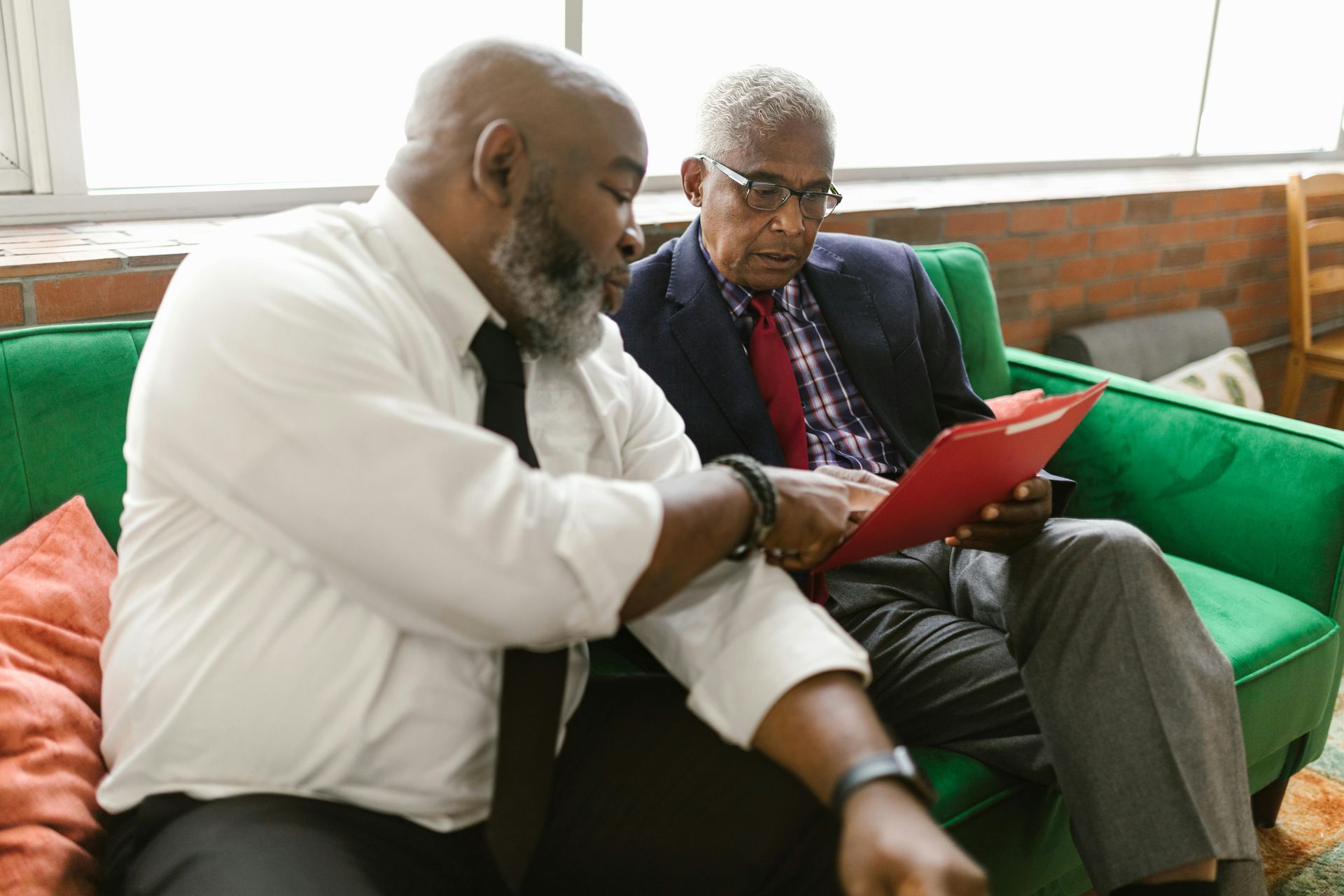 Two men seated on green sofa, reviewing documents in a red folder. One points at the folder.