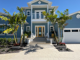 A blue two-story house with a light yellow front door and a brick walkway. Palm trees flank the entrance.