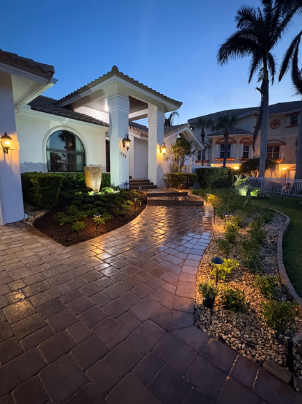 Night view of a well-lit home with a brick pathway. Landscaping with small shrubs and lights flanks the path.