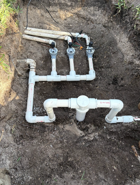 Irrigation system with three valves and a filter, made of white PVC pipes, installed in a dirt trench.