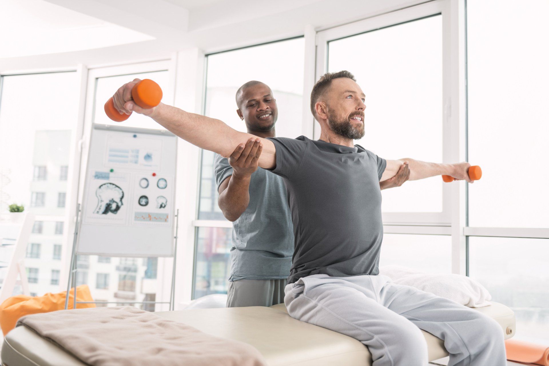 Man doing shoulder exercises with a therapist's help. Bright room, dumbbells.