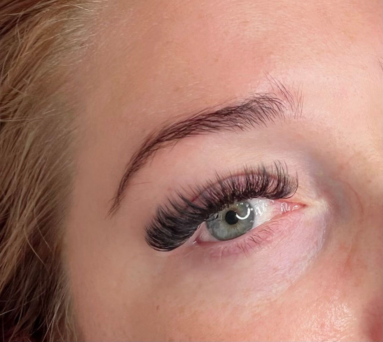 Close-up of eye with dark, thick eyelash extensions and styled, dark eyebrows. Light skin and a blue-green eye.