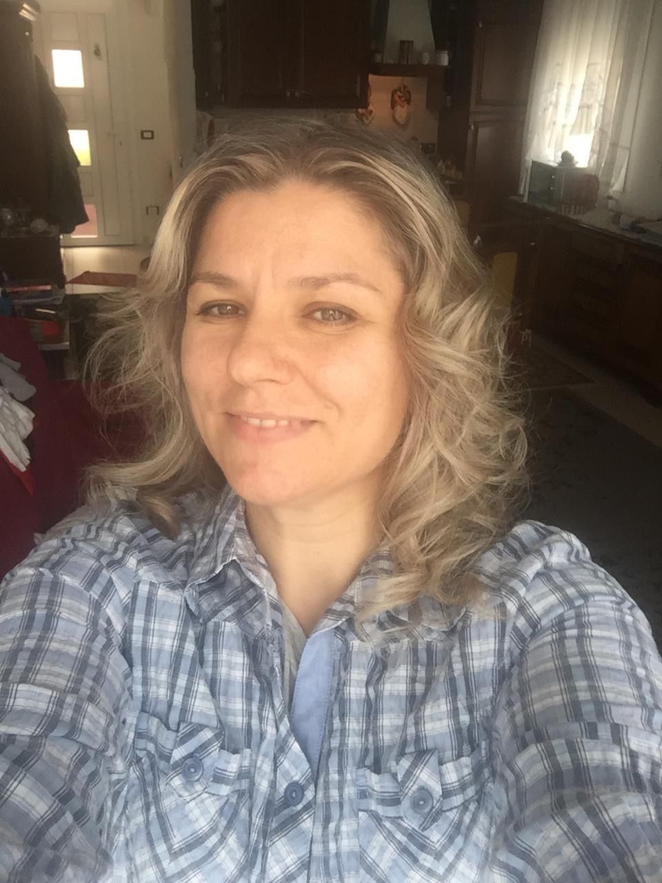 Woman with curly blonde hair in a blue plaid shirt smiles at the camera, inside a room.