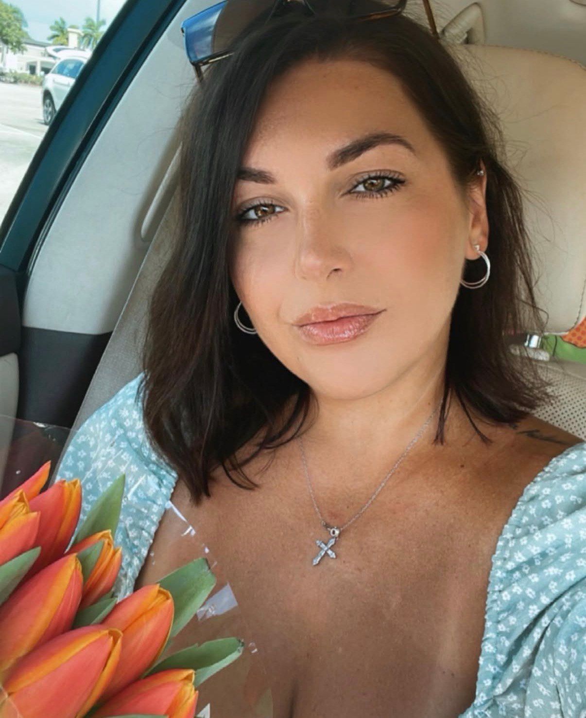 Woman in a car holding orange tulips, wearing a blue floral top, hoop earrings, and a pendant.