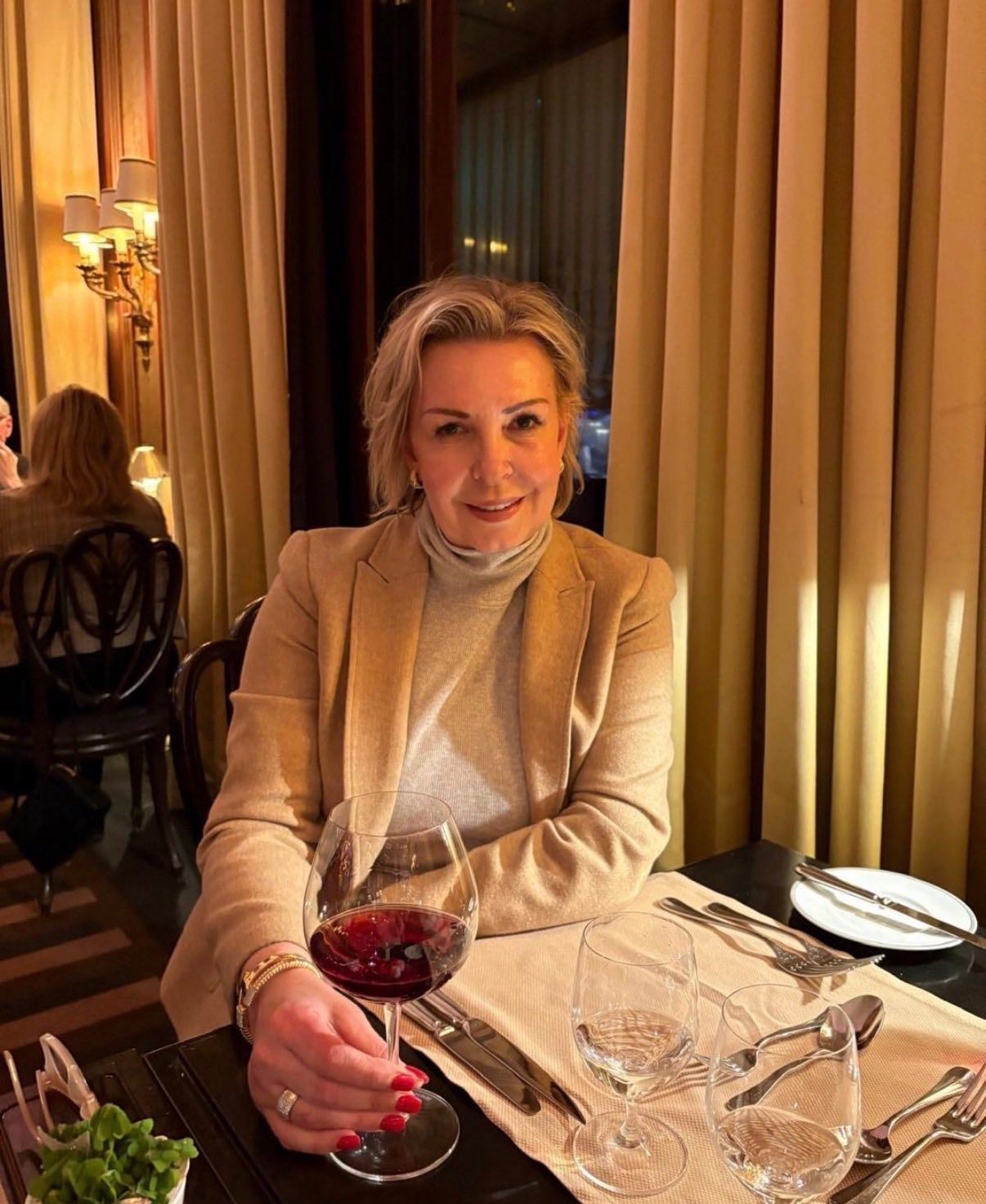 Woman seated at a restaurant table, holding a glass of red wine, wearing a blazer. Warm-toned lighting.