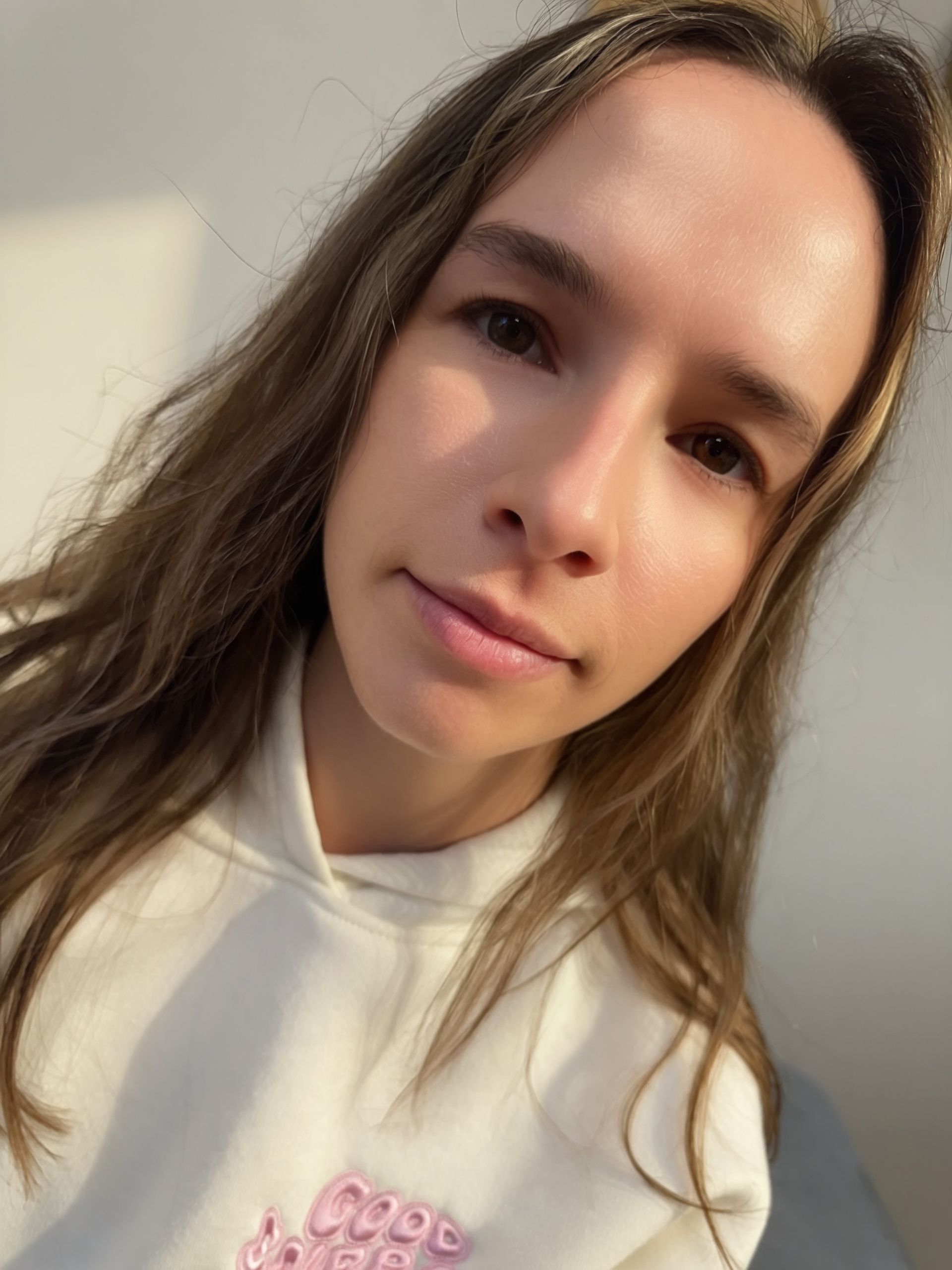 Woman with long wavy brown hair, wearing a white hoodie, looking at the camera with a slight smile.