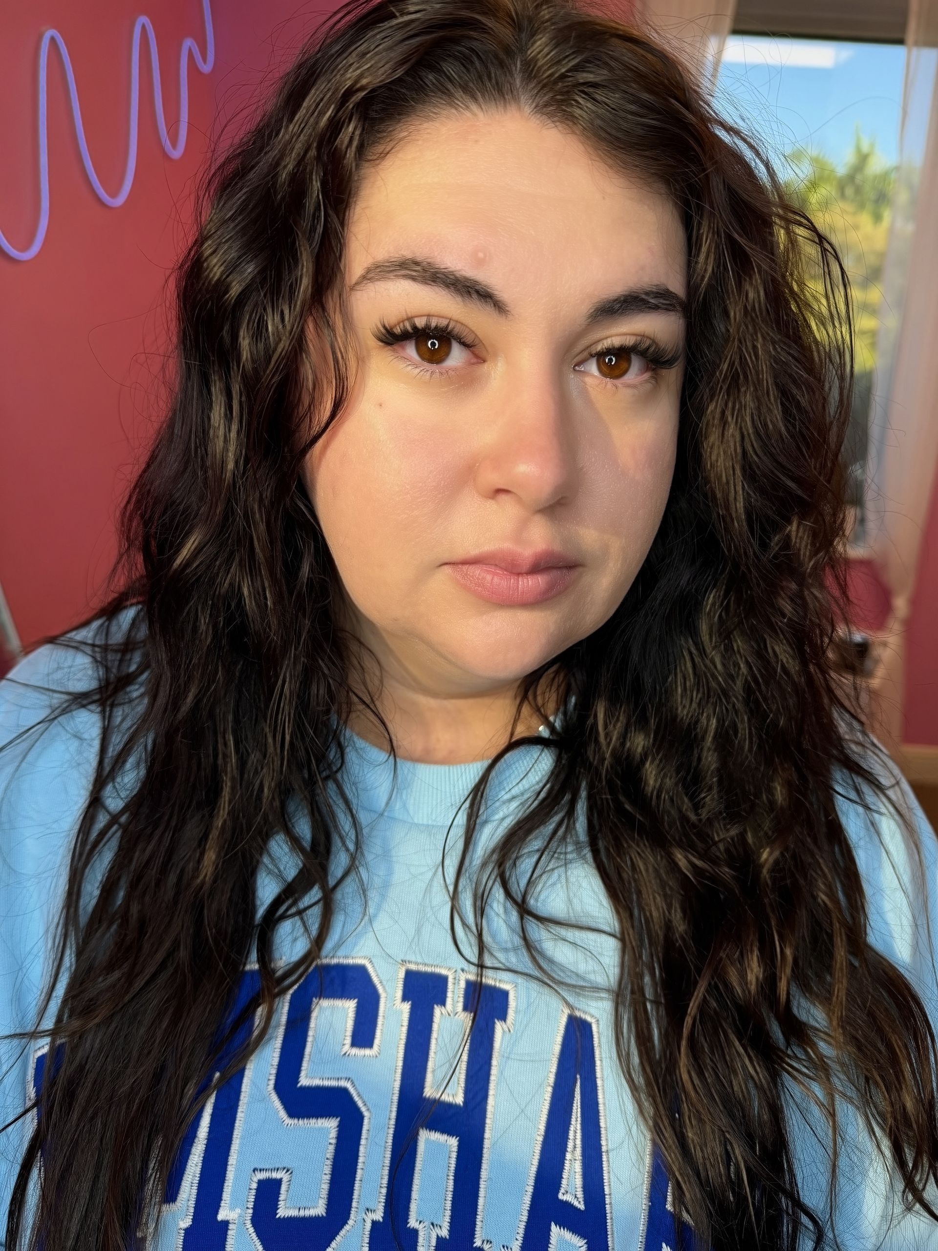 Woman with long dark curly hair, wearing a light blue sweatshirt, looking directly at the camera.