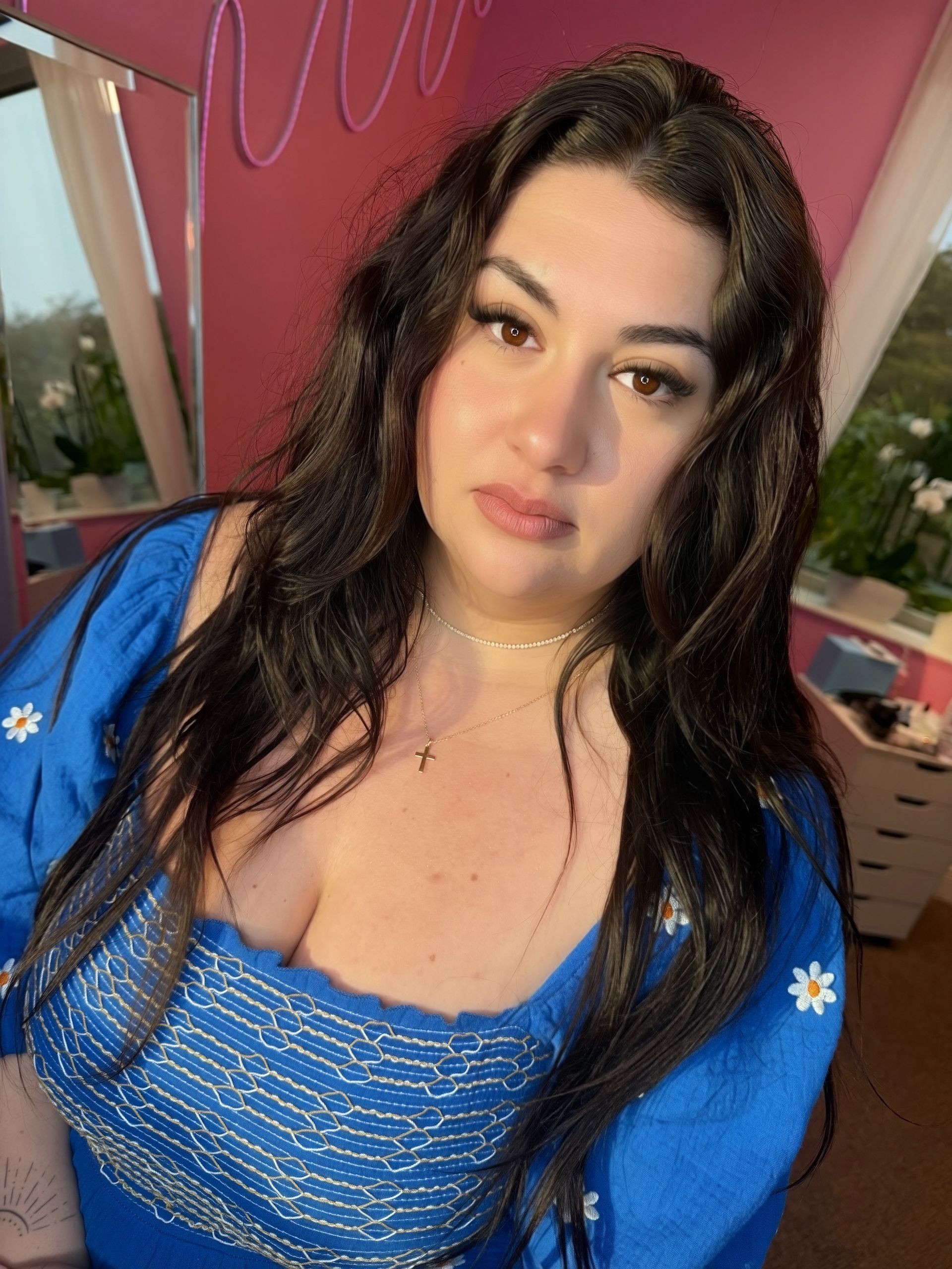 Woman with long dark hair wearing a blue daisy-embroidered top; gazing at the camera, pink background.