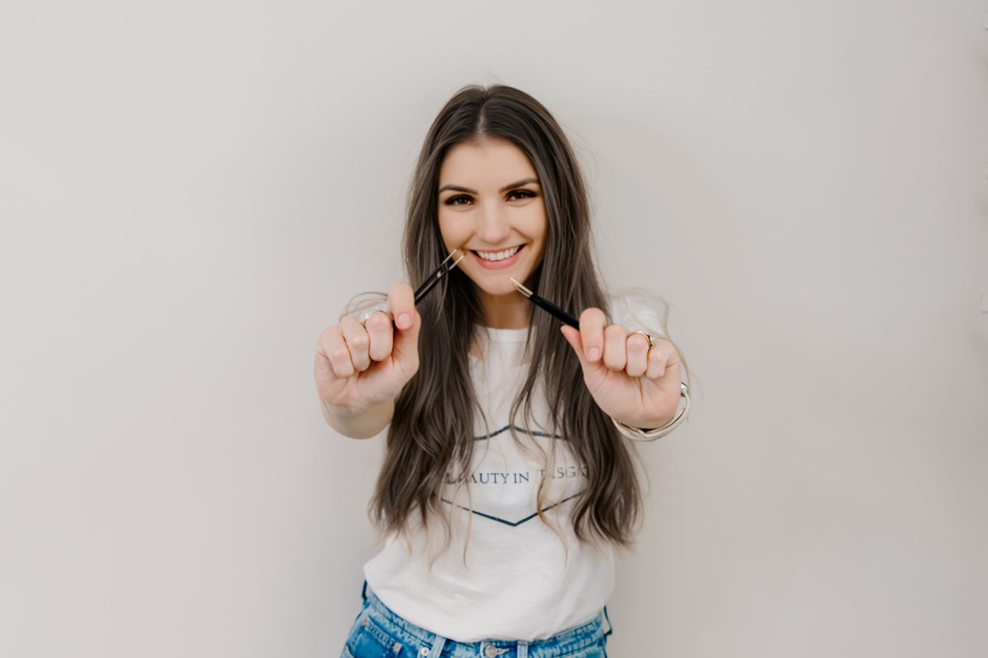 Woman smiling, holding pens, facing camera; white background.