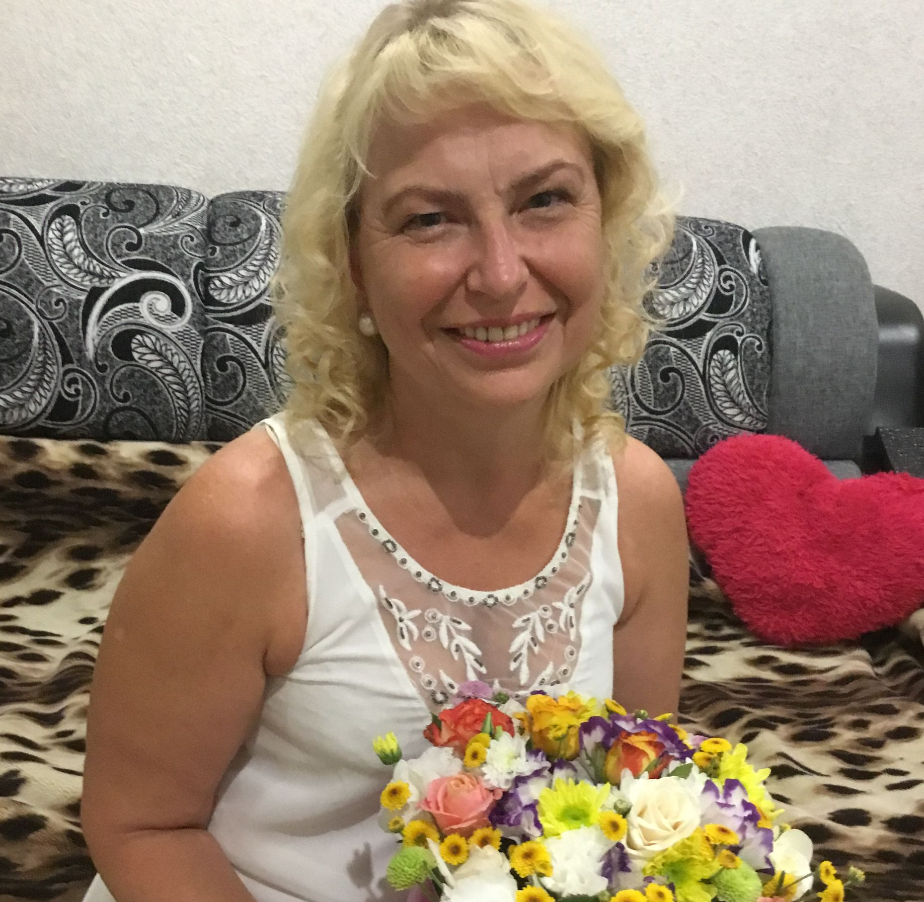 Woman holding flowers, smiling, sitting on a couch with a heart pillow, wearing a white top.
