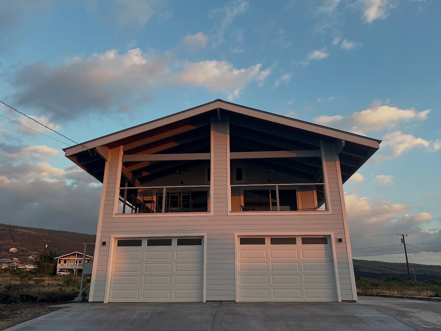 Two-story light gray house with two garage doors and a balcony under a partly cloudy sky at sunset.
