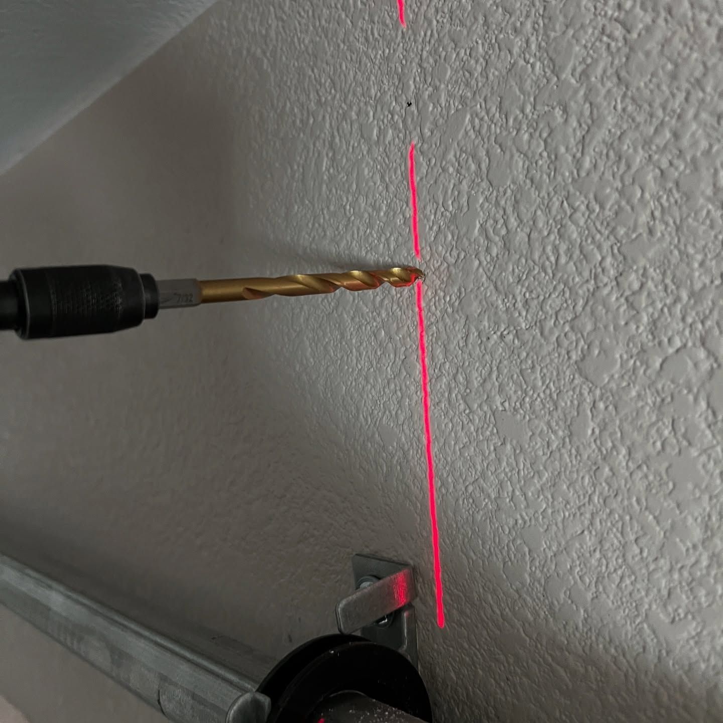 A drill bit against a textured wall, guided by a red laser line. A garage door track is at the bottom.