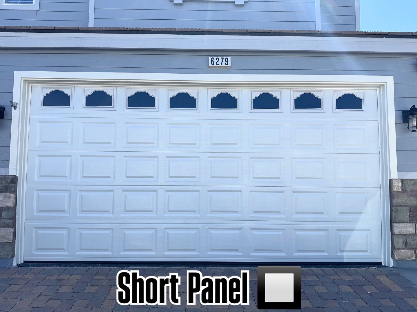 White short-panel garage door with arched glass panels, set in a house with stone accents, and a brick driveway.
