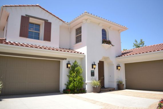 White two-story house with brown tile roof and garage doors. Includes small tree and outdoor lighting.