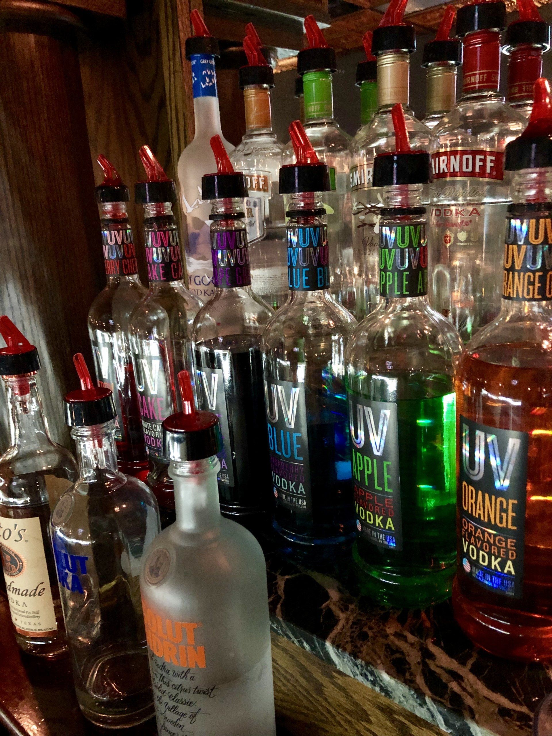 Many bottles of liquor are lined up on a shelf including uv vodka