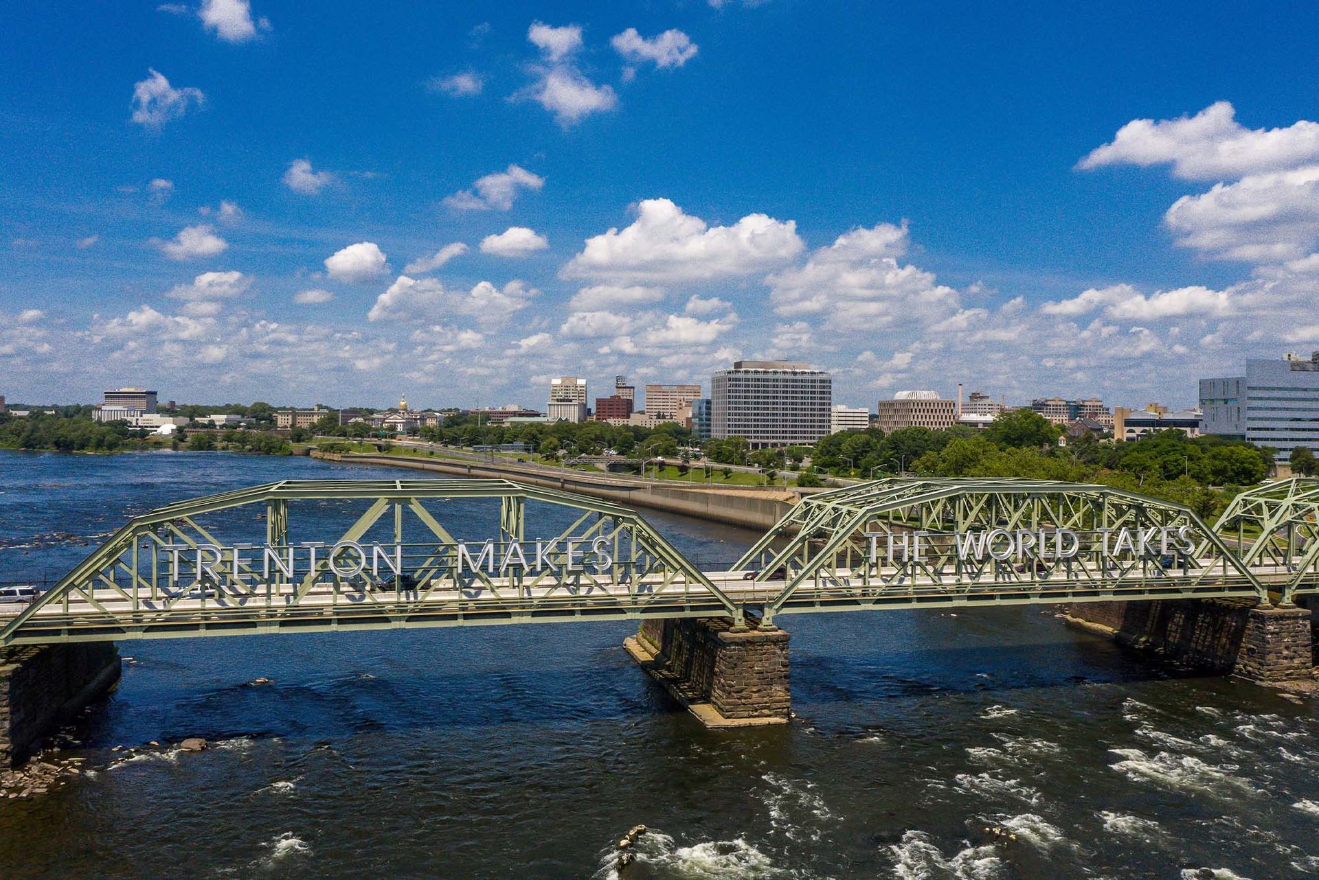 Wide river with a green metal truss bridge and city skyline under a blue, cloud-filled sky.