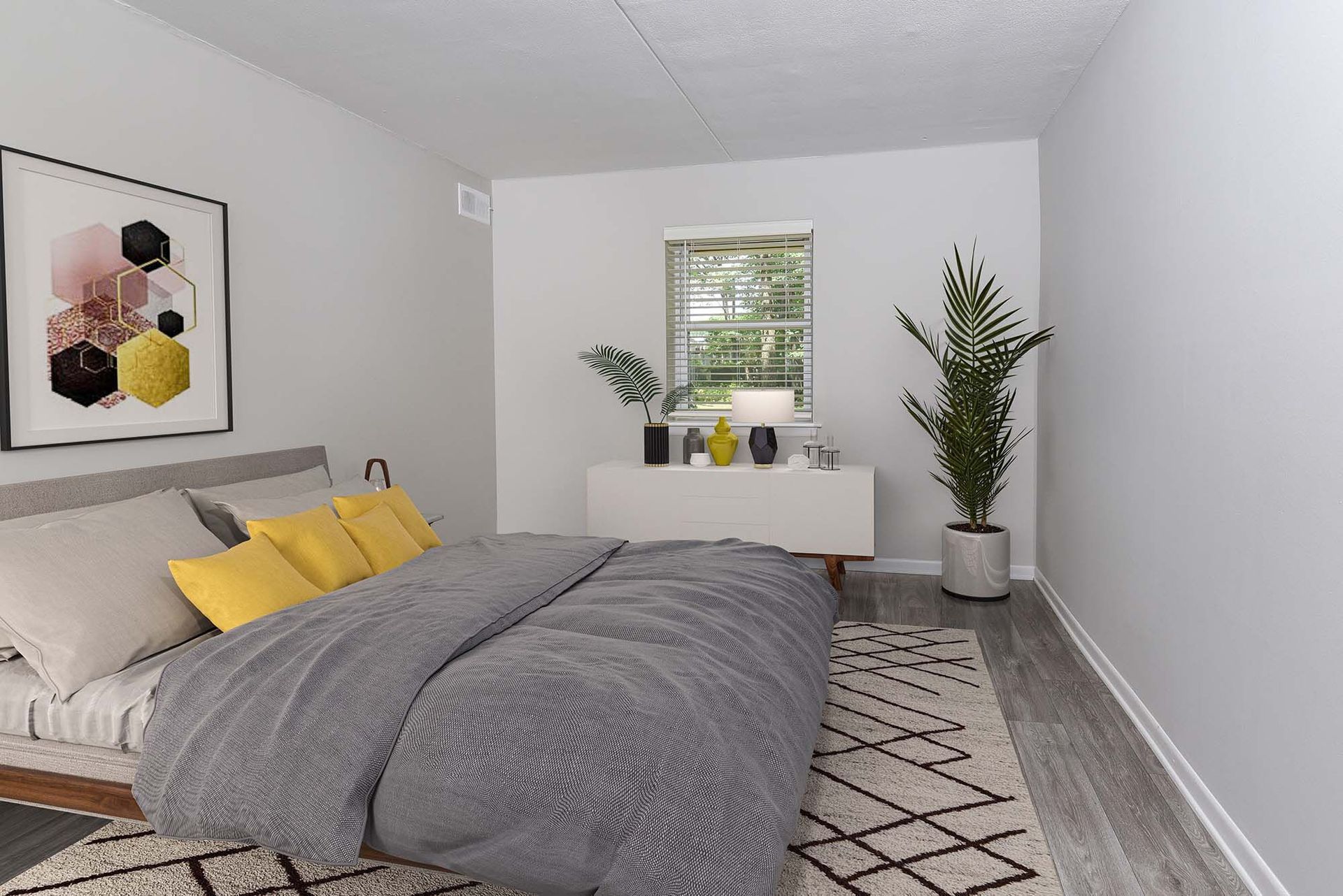 Bedroom in a modern apartment with a gray bed, yellow accent pillows, white dresser, and a tall plant.
