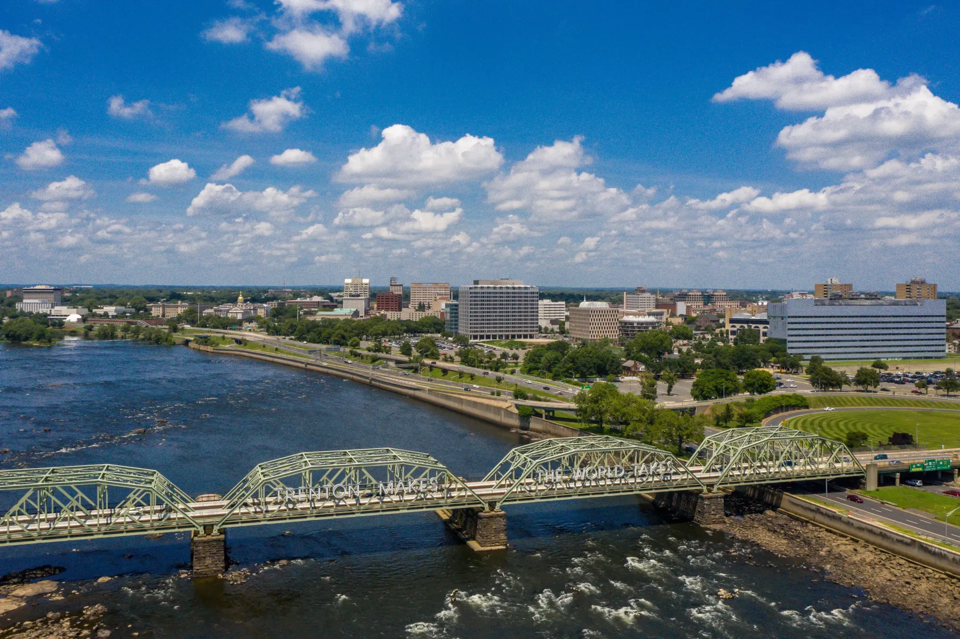 Aerial cityscape with a river, a steel bridge, and downtown buildings under a blue sky.