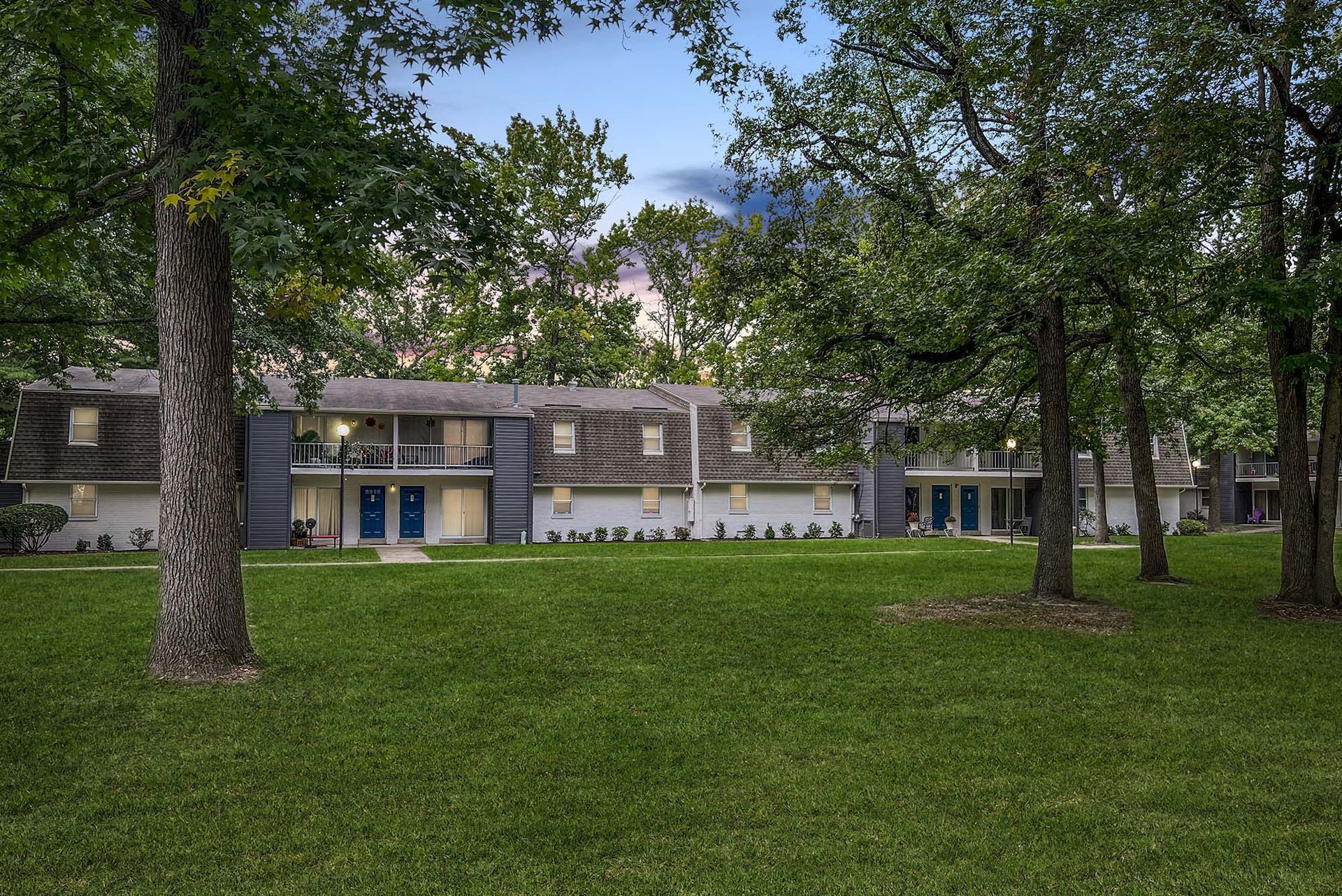 Exterior view of a two-story apartment building with blue doors, a green lawn, and surrounding trees.