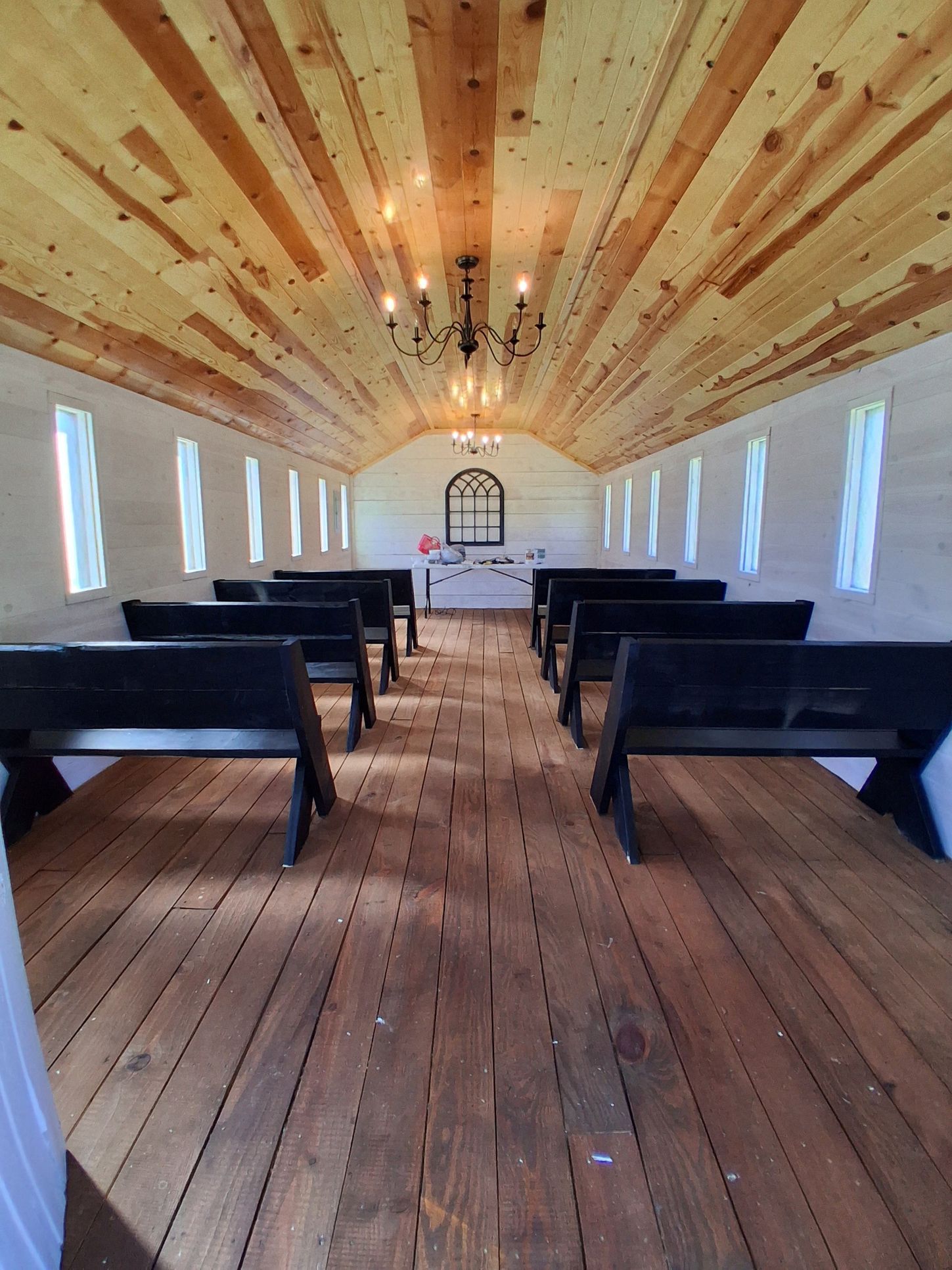 Interior view of a rustic chapel with rows of dark wooden pews facing an altar under a high, vaulted wooden ceiling.