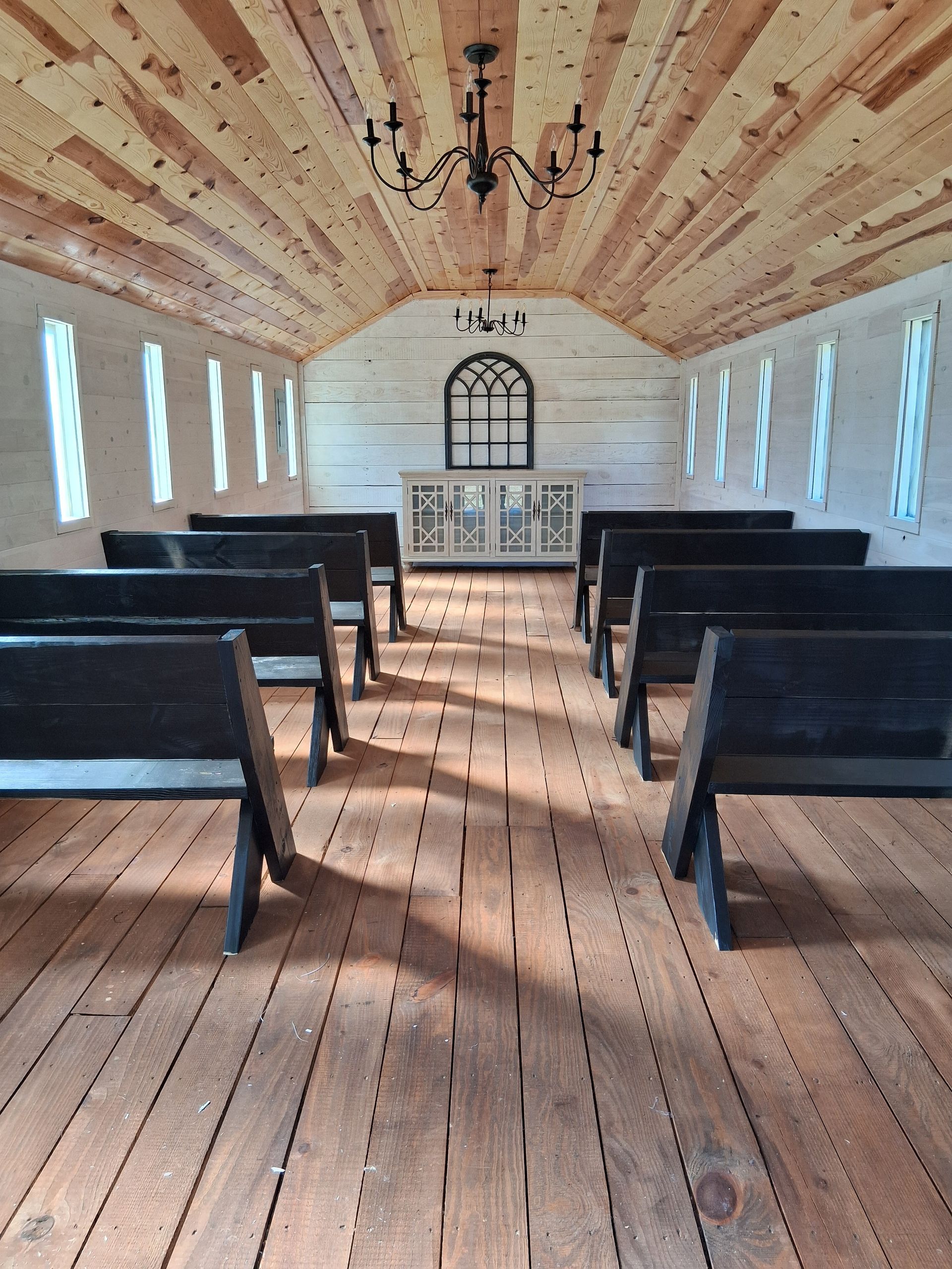 Interior of a rustic chapel with white walls, a wood-paneled ceiling, black pews, and a decorative arched wall window. Interior of a rustic chapel with white walls, a wood-paneled ceiling, black pews, and a decorative arched wall window.